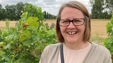NHS Sussex Woman smiles at the camera, wearing glasses with green fields in the background