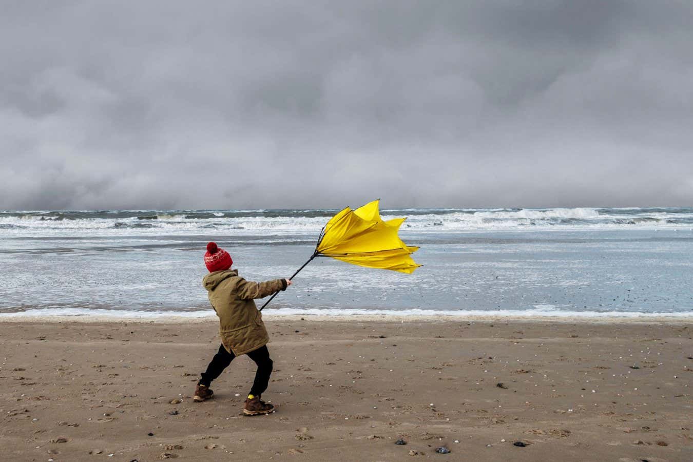 2DF6M9A Young boy struggling with large yellow umbrella on beach in stormy weather. Winter beach scene.
