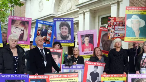 PA The families of those who have died while in the care of mental health services in Essex stand outside the inquiry protesting. There are around 20 people in the image which shows them standing with placards held against them and large posters of their loved ones. They show a range of faces, both men and women and young and old. One reads