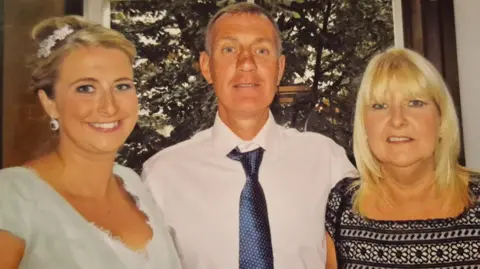 Family handout Three people stand smiling for the camera in front of a window, which shows large trees outside. To the left is a  young woman in a green dress. Her blonde hair is tied up in a bun with a floral display. She has white earrings dangling. A man in a white shirt and blue tie is in the centre with an arm around the two women. He has dark hair, which is turning grey. To the right is an older woman with blonde hair which hangs by her shoulders. She is wearing a black pattered top, which appears to have flowers embroidered into the pattern. 