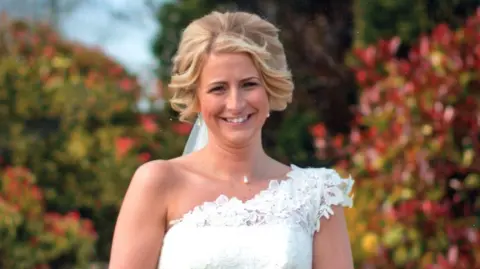 Quinn family photograph A woman in a white wedding dress smiles at the camera. She is holding a bouquet of white roses with both hands in front of her. Her blonde hair has been styled into curls, and a veil hangs from it behind her. The bride is pictured in a garden with a bench, medium level wall and bright bushes visible behind her.  