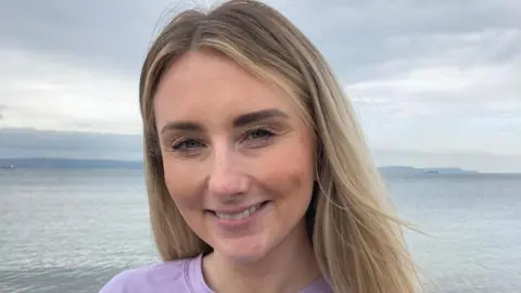 Shelley smiles at the camera as she stands on a beach. Her long hair, which is naturally dark with blonde highlights, drapes down behind her shoulders. Shelley is wearing a light purple t-shirt which reads 'wellness' and 'self love'. Behind her a small piece of sand is visible along with the sea, a cloudy sky and some mountains in the distance. 