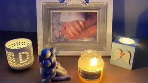 A photo of a stillborn baby's hand in a woman's hand. Beside it is a tealight holder with the letter D on it, a mother and baby small statue, a Yankee candle, and a blue candle with white angel wings on it.