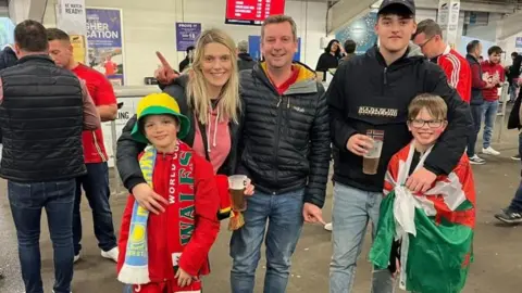 Family photo Elen is standing with her family in the concession area of a sport stadium. She is standing close to a man and a teenage boy in black jackets. Two boys are with them one wearing a wales flag and the other has a Wales footbal scarf 