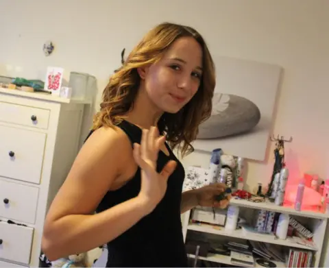 Family handout Alice is standing in her bedroom on her 18th birthday. She is wearing a black sleeveless dress and is holding her hand up as if she is waving. There is a shelf and chest of drawers behind her and an artwork on the wall.