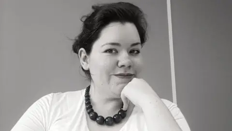 Rachael Clarke Rachael, who has dark hair, wearing a white t-shirt and a chunky bead necklace looks into the camera while resting her chin on her hand.