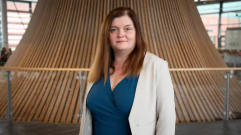Sioned Williams, who has brown, straight hair, looks directly into the camera wearing a cream jacket and blue wrap dress.