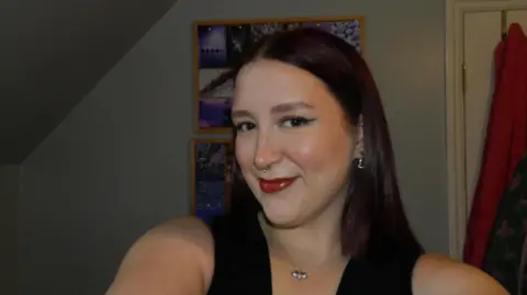 Tilly Short Selfie photograph of Tilly Short who smiles at the camera. She has dark brown hair, and wears red lipstick. She has a silver necklace on and a silver nose ring.