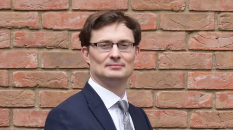 Steve Copley Tom Quinn wears a dark navy suit, a white shirt and a paisley print tie. He wears black rectangle glasses and has brown short hair. He stands infront of a red brick wall.