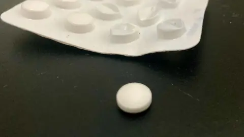 A single white aspirin tablet next to a packet.   