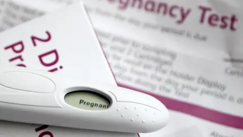 PA A positive pregnancy test lies on top of the test box which is white with magenta writing, the instructions for the kit are blurred out in the background.