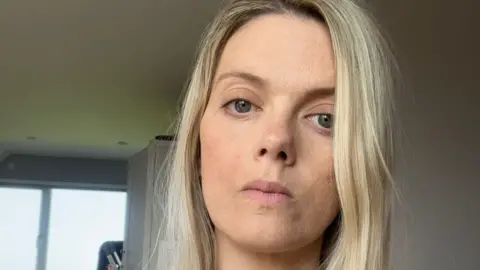 Elen Hughes A selfie of Elen Hughes inside a house with light grey walls and a window visible behind her. She looks serious and has long blonde hair and there is a light grey cupboard behind her