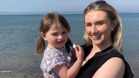 BBC Shelley and Ruby smile as they stand on the shoreline at the beach looking at the camera. Ruby is in Shelley's arms and is eating a marshmallow. She has brown hair in a double ponytail and is wearing a white t-shirt with colourful patterns on it.  Shelley has naturally dark hair which has been dyed blonde. She has sunglasses propped on top of her head and is wearing a black shirt. Shallow water is visible behind them, which stretches out to the mountains on the horizon. 