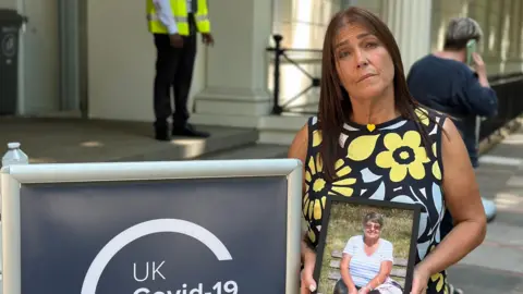 Getty Images Nicky Hastie, in a floral summer dress, standing outside the Covid inquiry, holding an image of her mother Margaret.