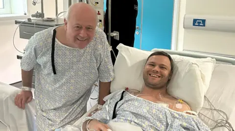 Kieran Innes Kieran is lying in a hospital bed and smiling. His dad Brian is standing beside the bed wearing a hospital gown. Brian is bald. Kieran has brown hair and a stubbly chin.