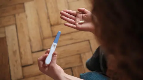 Getty Images A woman holds a pregnancy test in one hand and a pill in the other