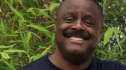 Handout Head and shoulders of a smiling Mel Tottoh. He is wearing a navy t-shirt and standing in front of green bamboo foliage