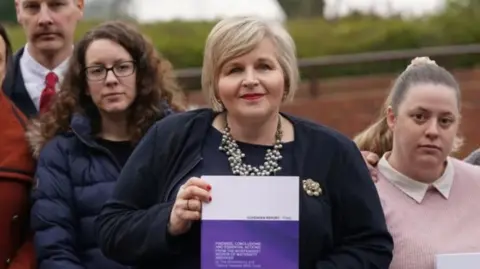 BBC A woman with bobbed blonde hair is wearing a chunky necklace, flower broach, black top and black cardigan. She is standing in front of a gathering of woman and is holding purple and white book in her hands that says report on it.