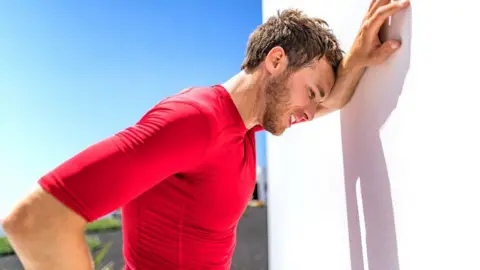 Getty Images A sweating man with brown hair and wearing a red top leans against a white wall in the sun because he is overheating.