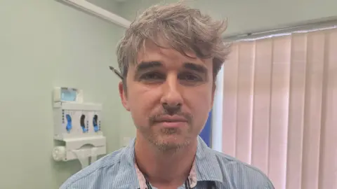 Dr Matthew Jones with short brown hair and a slightly greying brown beard. He has a biro ball point pen tucked behind his ear and is looking directly at the camera. He has a blue shirt with small white stripes and the background is lime green walls, with a dispenser for glove and waste bags. There is also a window covered by cream blinds.