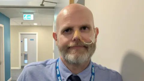Gareth, a bald, white man with a thick greying beard leaning on a white wall. He is looking into the camera with a neutral look on his face. He is wearing a light blue shirt with a navy blue tie, and a blue NHS lanyard. He has a tube that goes into his left nostril with plasters holding it in place. The background includes a hallway with white and blue walls, white doors and a neon green fire escape sign.