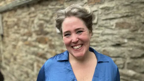 BBC A woman looking at the camera and smiling. She has short hair. She is wearing a blue top. There is a blurred brick wall behind her.