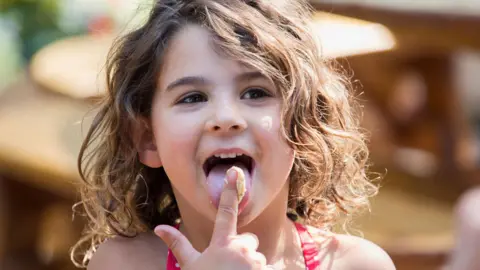 Getty Images A young girl with wavy brown hair, wearing a top with red straps, licks her index finger, which has food on it, in the outdoors with the sun shining behind her