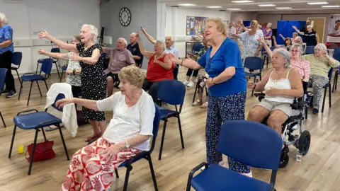 BBC Men and women are taking part in an exercise class in a hall. Some are standing and some are seated and one woman is in a wheelchair. They are following the instructor's arm movement. The hall is nearly full and there is a clock on the wall, with a noticeboard.