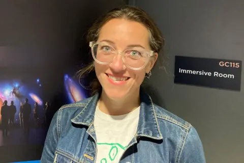 Kate Cox wearing a denim jacket and translucent glasses, smiling at the camera. She is in the new campus building, standing in front of a door that has a sign reading “Immersive Room