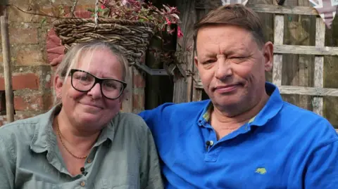 JOHN FAIRHALL/BBC Tony Henderson with his wife Sarah sitting outside their house in Bacton in Norfolk. He is wearing a royal blue polo shirt and he has his arm around his wife's shoulders who is wearing an olive green shirt. 