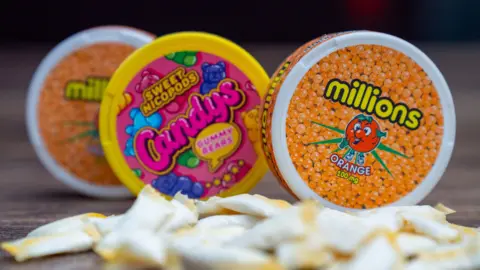 BBC A photo of three small tubs of nicotine pouches. One is orange and has a picture of a smiling orange on the cover with loads of small oranges under neath the word Millions. It does not mention the word nicotine. The Candys pack is pink with colourful gummy bears on it. It says