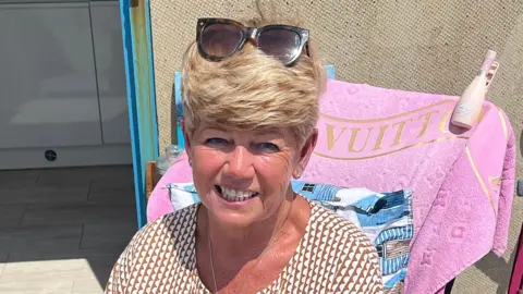 BBC Debbie Booker is sitting outdoors. She has short blonde hair and is wearing sunglasses on top of her head and a patterned shirt. Behind her, a pink towel with the word