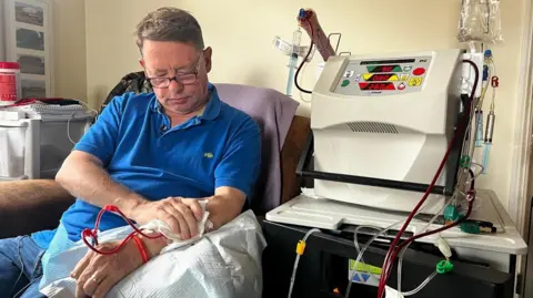 JOHN FAIRHALL/BBC Tony Henderson sitting in his living room undergoing home dialysis. His dialysis machine is to his left and you can see tubes running into his left arm. You can see him applying pressure to his left arm with his right arm at the fistula site. Mr Henderson is wearing a royal blue polo shirt and has glasses on.