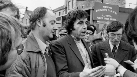 Getty Images A black and white shot of a man speaking to journalists - who are out of the shot. He is surrounded by other men and is standing in front of a sign which reads