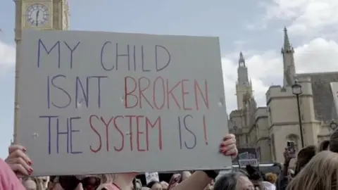 A sign being held up amongst a crowd in front of the Palace of Westminster. Big Ben can be seen in the background.
The sign reads: My child isn't broken the system is!