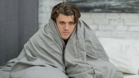Getty Images A young man with tousled brown hair is looking glum and a bit ill, wrapped up in a grey-coloured duvet while sitting on his bed