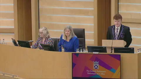 Scottish Parliament A teenage boy standing up behind a podium in a parliamentary building, wearing a black school uniform with purple trim and tartan tie. Two women are sitting along from him, one wearing a blue suit and the other in a multi-coloured top. The walls and decor of the room are beige.