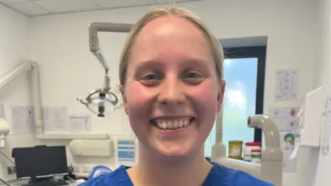 A young woman is wearing blue dental scrubs and has a University of Plymouth name badge on that says