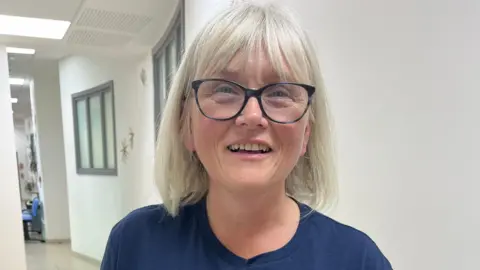 A woman with black glasses and a grey bob haircut is smiling to the camera. She is wearing a blue T-shirt with