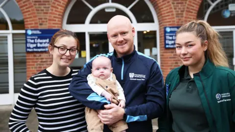 University of Worcester Two women standing either side of a man who is holding a baby. On the left is the baby's mother, who has ginger hair tied back and is wearing glasses and a black and white striped top. The man, the baby's father, is bald with ginger facial hair and is wearing a navy top that says 