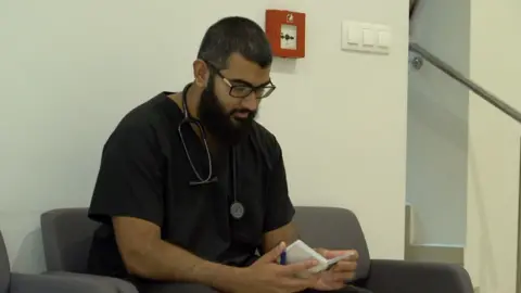 Mohammed is wearing medical scrubs and is sat in a waiting room chair reading a notepad