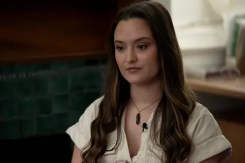A young woman with long brown hair is pictured face on however she is looking down to the lower left of the screen. She has a short-sleeved white shirt on and a necklace. 