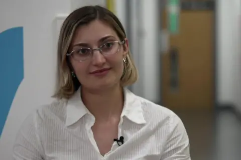 A woman with shoulder-length blonde hair is pictured facing the camera but her eyes are looking slightly to the right. She has a white shirt on. In the background is a corridor of a clinic so white walls and a brown fire exit door.