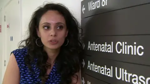 On the left is a woman wearing a sleeveless dark top with a blue pattern. She has long black hair and big hoop earrings. She is standing next to a sign saying 'Antenatal Clinic'