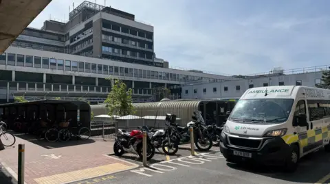 Steve Hubbard/BBC The exterior of Addenbrooke's Hospital, a mix of white and grey low-leve and multi-storey buildings, with a parked ambulance and motorcycles in the foreground next to a covered bicycle rack.