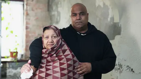 Bushra and Atif standing close together indoors against a wall with peeling paint and visible damage. Atif is wearing a dark hooded sweatshirt and has an arm around Bushra, who is wrapped in a patterned shawl with red and beige triangles. A window with a potted plant is partially visible in the background.