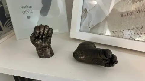 A small set of dark grey castings were made of Mabel's hands and feet. They are placed in a small white cabinet, in front of various other items to remember Mabel such as a printed footprint of her and a framed picture of her.