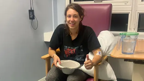 Kat Denisi Kat is wearing a black t-shirt and is sitting in a chair. She is holding a cardboard sick bowl and has brown hair.