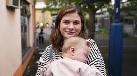 A portrait of Holly and her young daughter Ettie. Holly is a young woman aged 30 with long brown hair and wearing a striped blue top. She is carrying her baby Elly who is asleep in her arms and wearing a very light pink top with long sleeves.