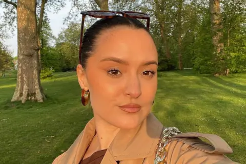 BBC A selfie of a young woman in a park. She has brown hair pulled back and tortoiseshell sunglasses on her head. She has a light brown mac on  and dark brown hooped earrings. Trees and grass are in the background.